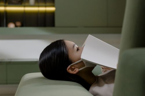 Free Side view of a woman lying down with a book and face mask in a modern interior. Stock Photo