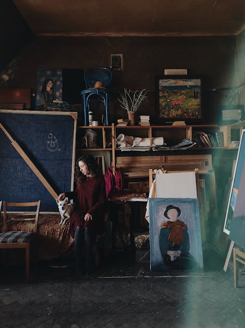 Full body of female artist sitting in modern art studio with abundance of creative pictures and painting tools on wooden shelves