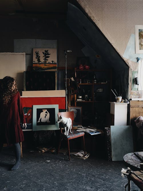 Anonymous female artist standing in creative workshop with abundance of pictures and artworks near instruments for painting in modern studio