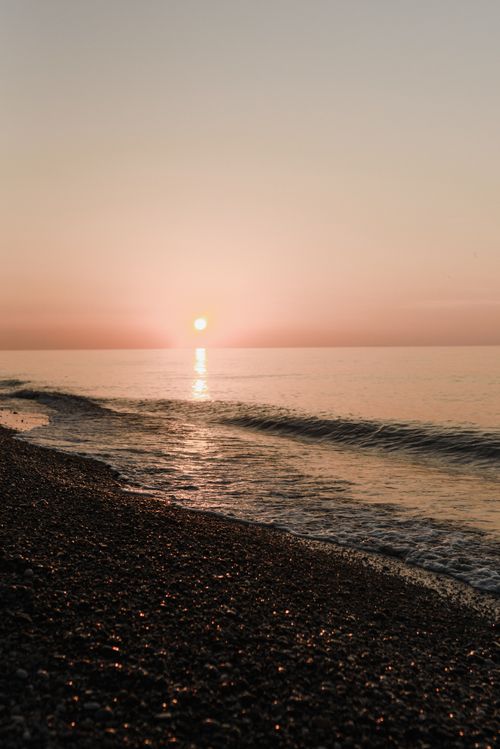 Free Peaceful beach scene with sunset reflecting on calm ocean waves and pebbled shore. Stock Photo