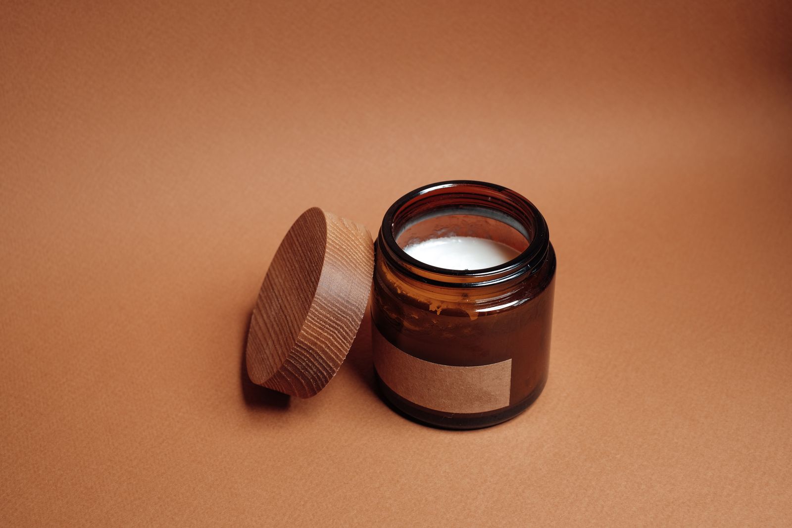 Open amber glass jar of white cream beside its wooden lid on a terracotta background