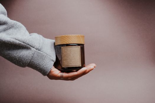 A close-up of a hand holding a glass jar with a wooden lid against a brown background, ideal for product designs.
