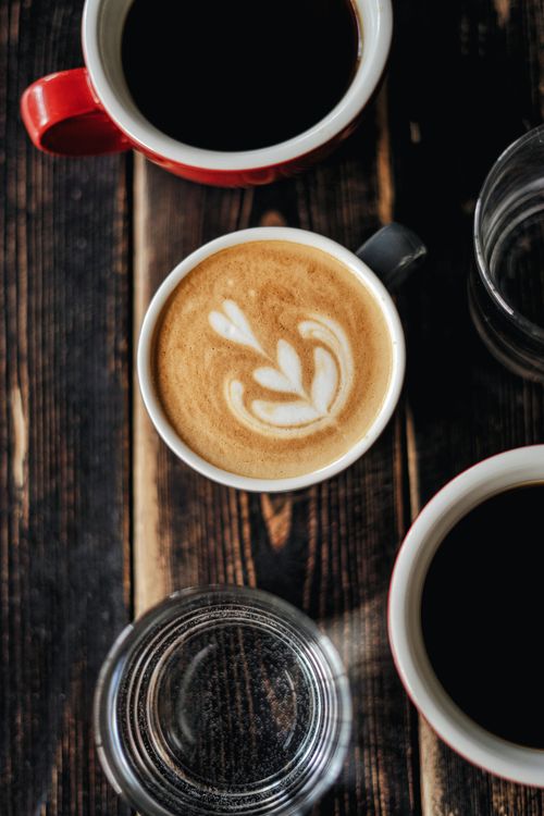 Free A beautifully crafted cappuccino with latte art surrounded by coffee cups on a rustic wooden table. Stock Photo