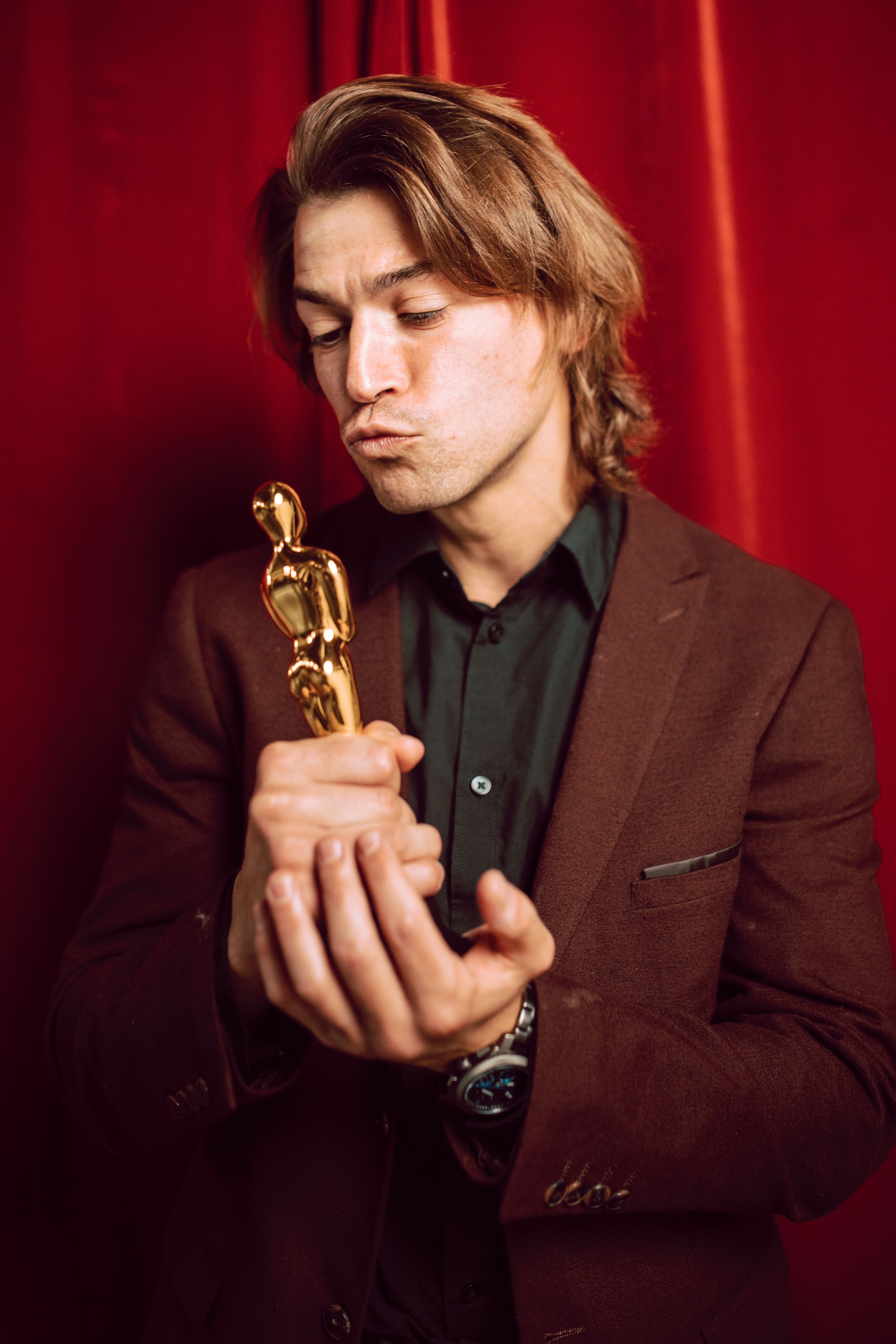 Caucasian man in suit holding a gold trophy, red curtain background.