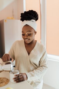 Smiling African American woman pours coffee from a moka pot into a cup in a sunlit kitchen.