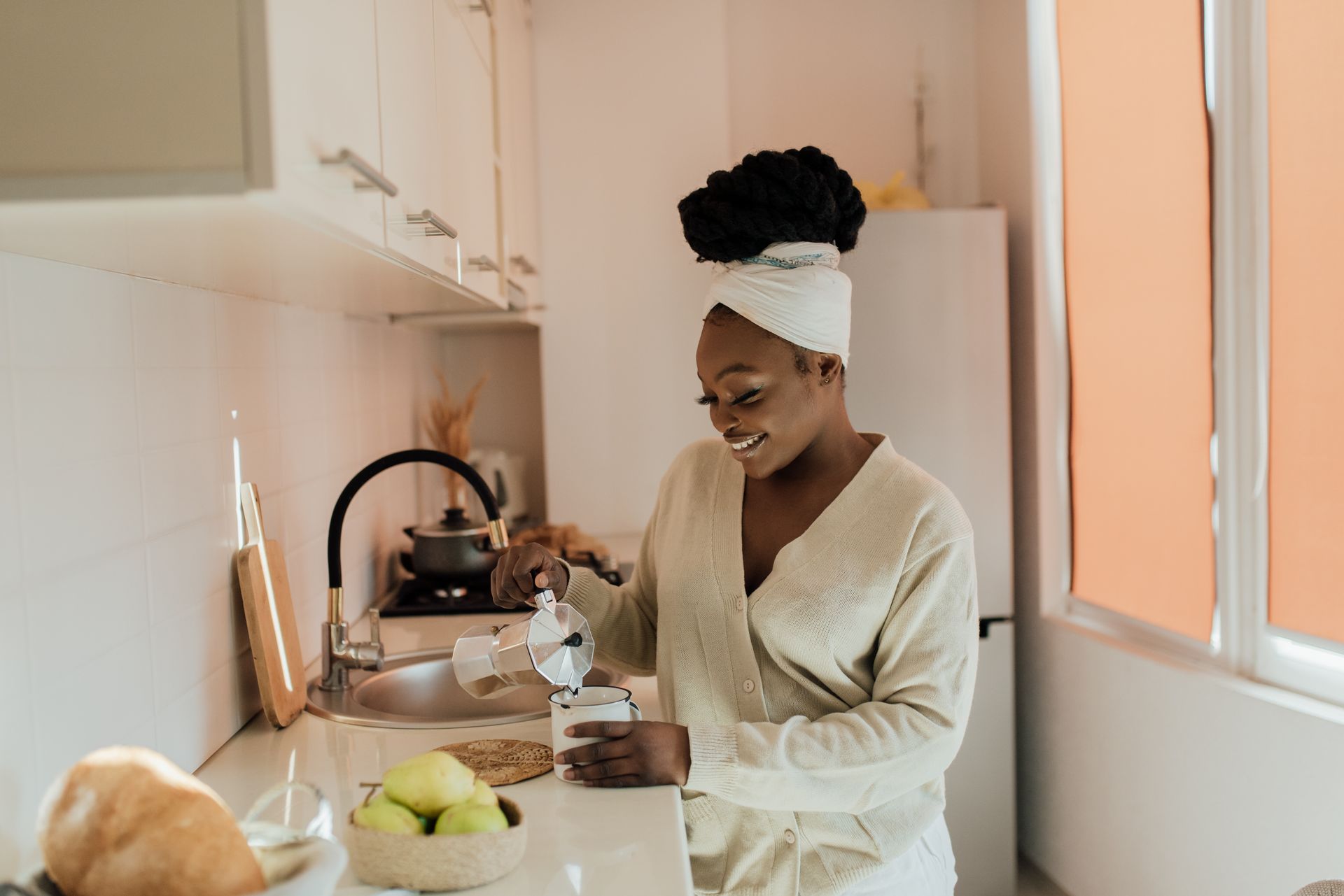 Smiling African American woman pours coffee using a moka pot in a bright kitchen setting.