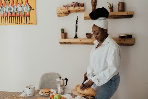 Free A woman slicing bread on a table in a cozy kitchen setting with art and decor. Stock Photo