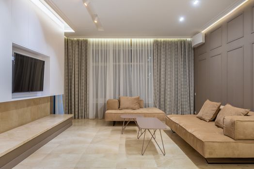 Comfortable big beige sofas with cushions and small coffee tables placed in spacious living rom in front of modern TV hanging on wall