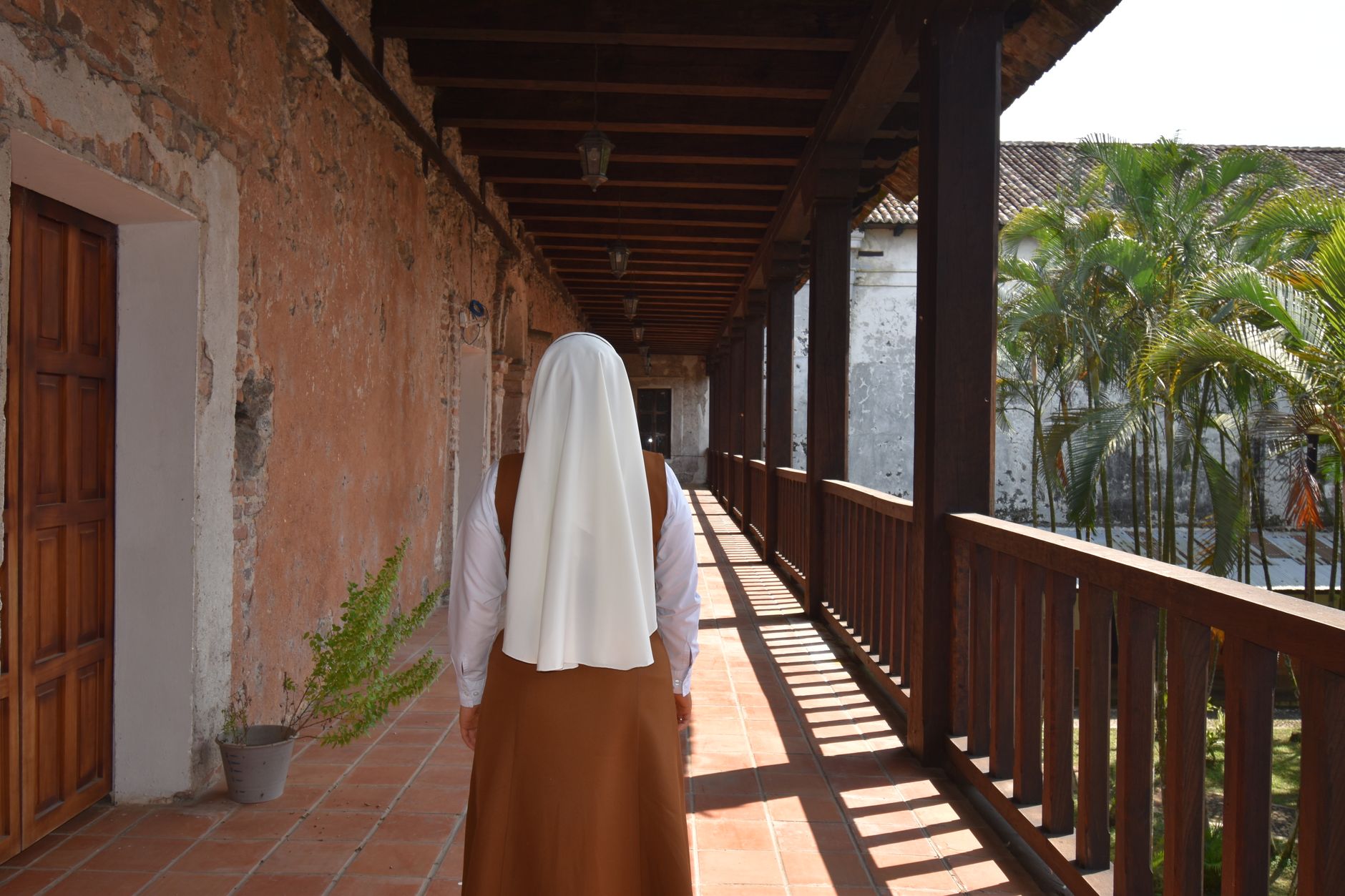 Rear view of a nun walking on a wooden balcony in a historic building under a bright day.