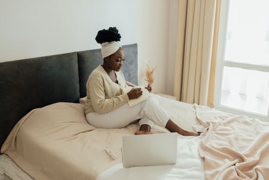 A woman with afro hair writing in a journal while seated on a bed next to a laptop in a cozy bedroom.