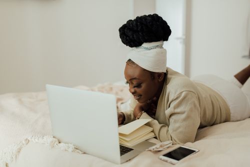 Free A woman with afro hair working on a laptop while lying on a bed in a cozy home setting. Stock Photo