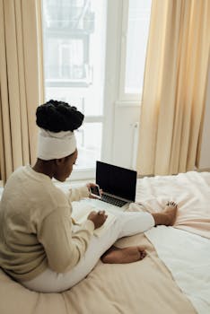 Woman working remotely, balancing gadgets and notes in a bright bedroom setting.