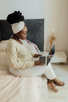 Black woman sitting on bed with laptop, embodying modern remote work lifestyle.