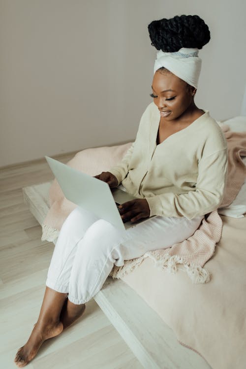 Woman in a White Sweater Sitting on a Bed while Using Her Laptop