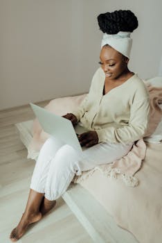 African woman with headwrap sitting on bed using laptop, working from home.