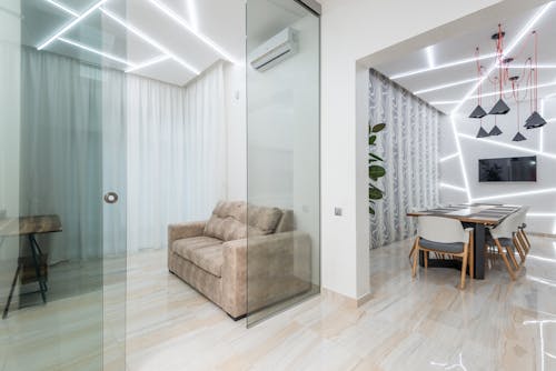 Light modern studio apartment with sofa behind glass walls and dining table under modern geometric illumination on ceiling