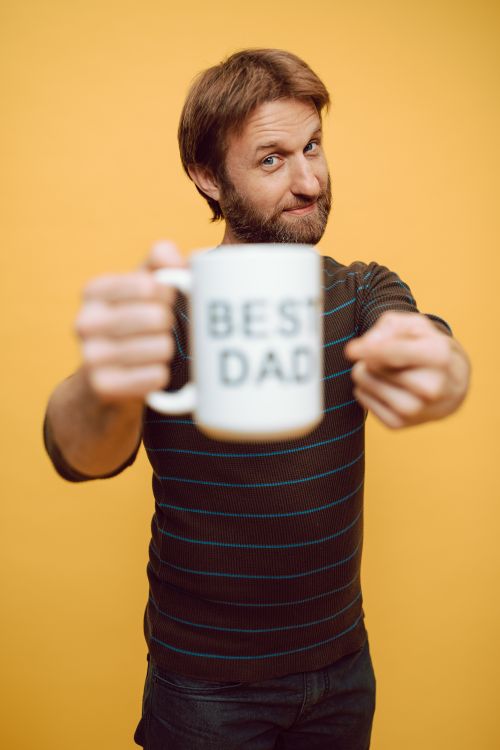 Free Cheerful bearded man holds a 'Best Dad' mug against a vibrant yellow backdrop, embracing fatherhood. Stock Photo