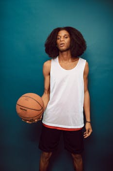 Athletic young man in sportswear holding a basketball, standing against a blue backdrop.