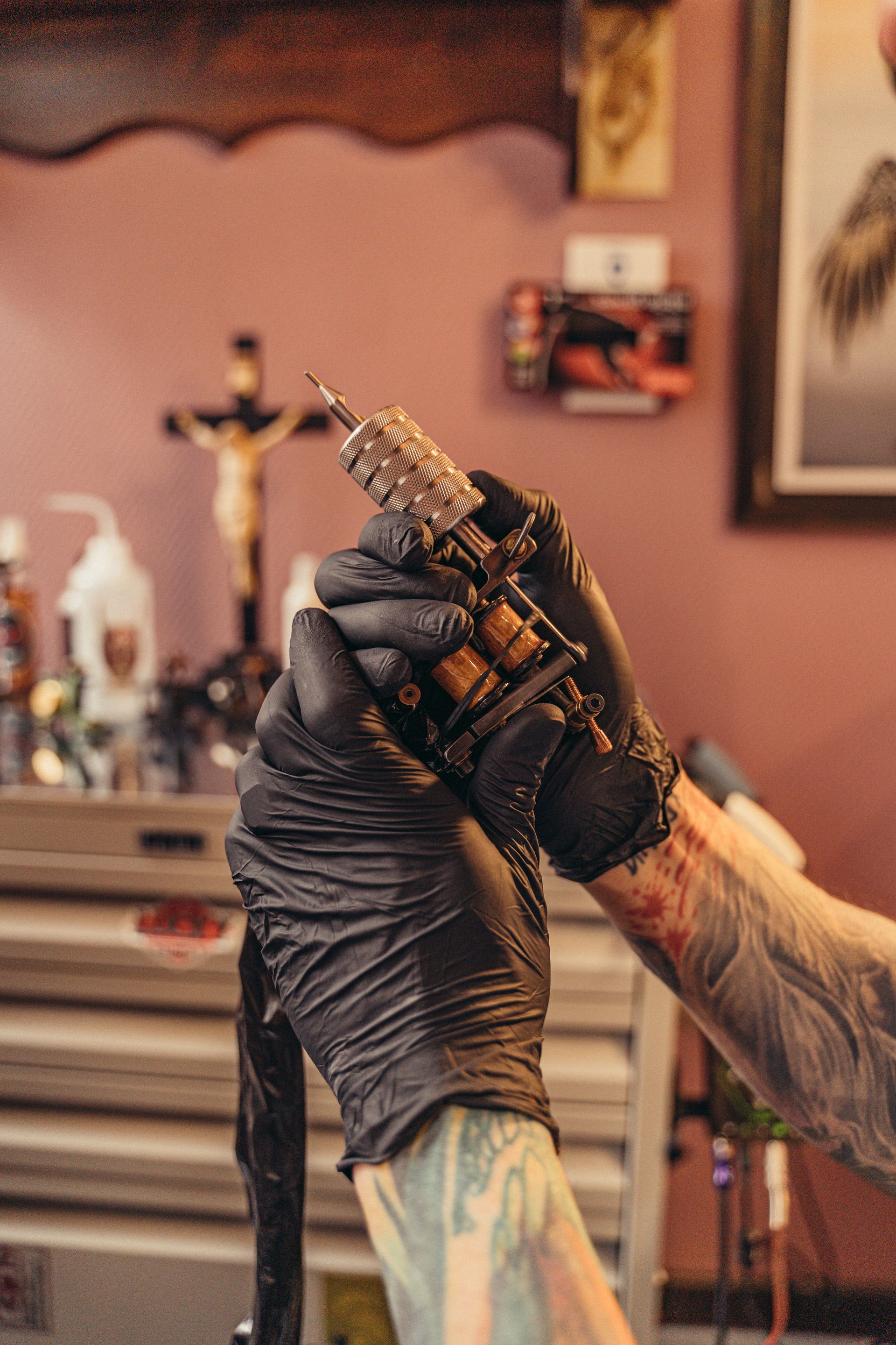 A tattoo artist wearing black gloves holds a tattoo machine in a studio, with tattooed skin and studio supplies visible in the background.