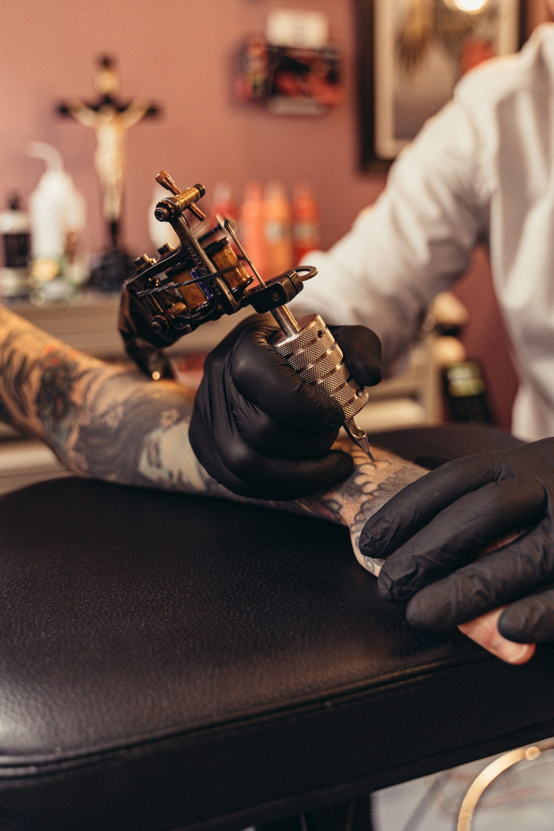 Close-up of gloved hands holding a tattoo machine over a tattooed client’s arm in a warm studio setting.