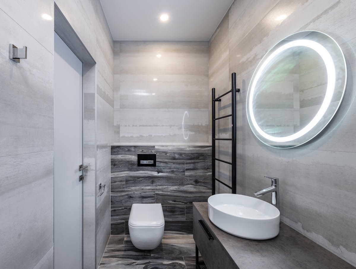Contemporary bathroom with toilet bowl and washstand between cabinet and oval shaped mirror with glowing lamp