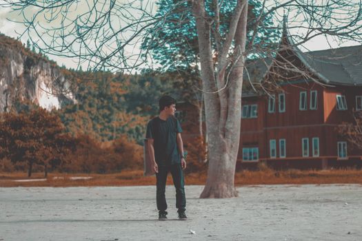 A young man stands on a sandy path in front of a rustic wooden house during autumn.