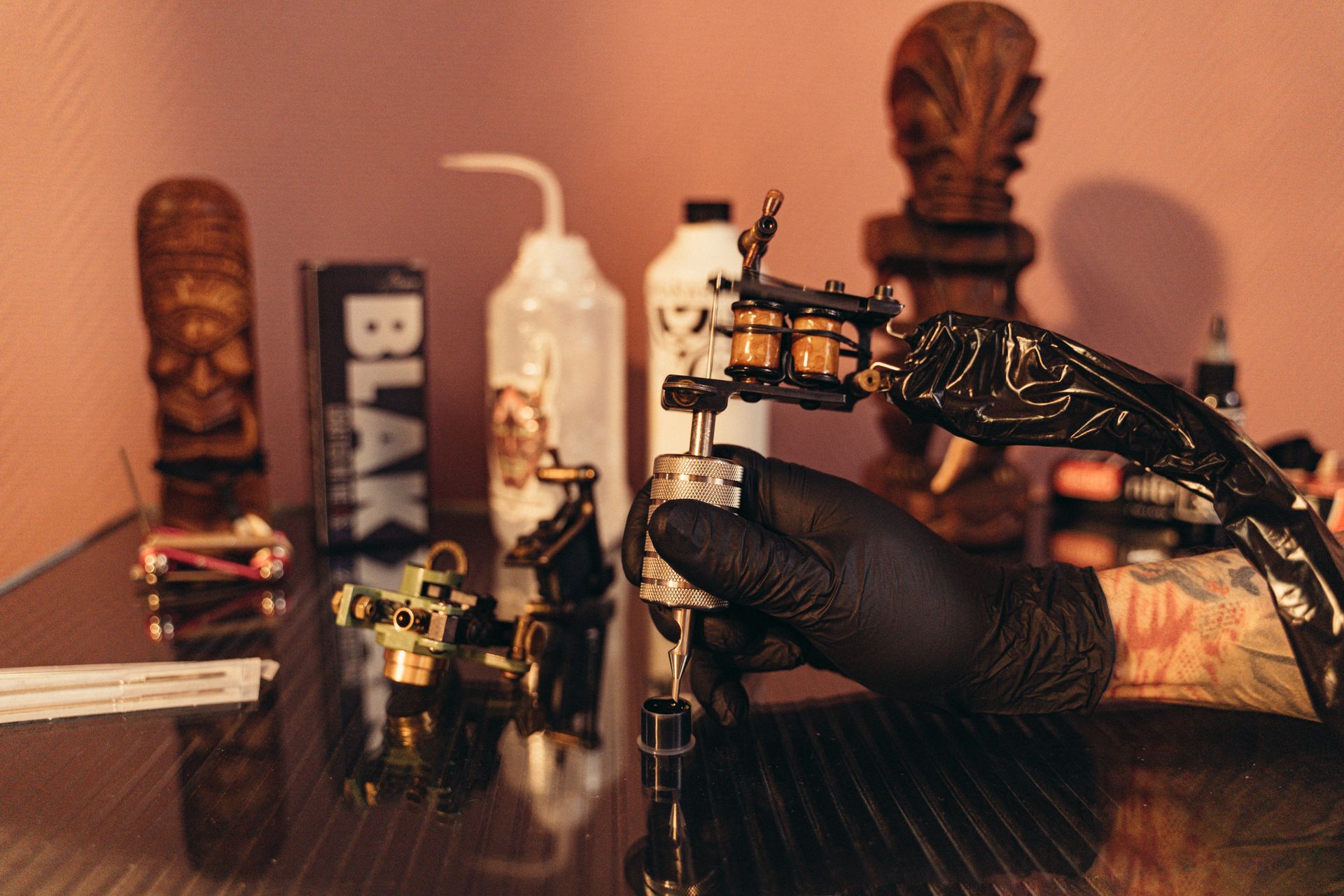 Black-gloved hands hold and adjust a tattoo machine in a studio with additional machines, bottles and equipment in the background.
