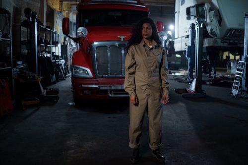 Free A woman in a uniform stands confidently in an automotive repair shop with trucks around. Stock Photo