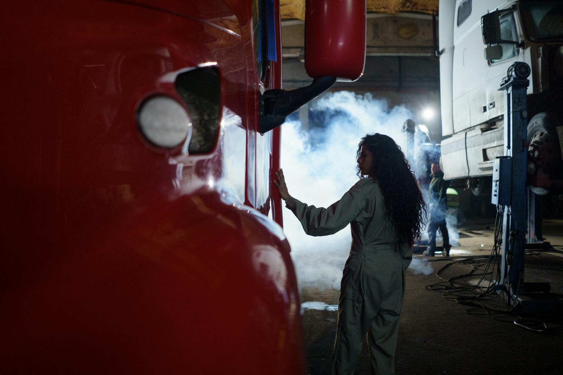 A female mechanic examines a truck in a dimly lit garage filled with smoke.