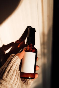 A hand in a cozy sweater holds an amber spray bottle with blank label in soft light.