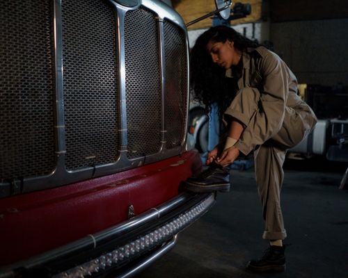 Free A woman mechanic in coveralls ties her shoes near a truck bumper in a garage setting. Stock Photo