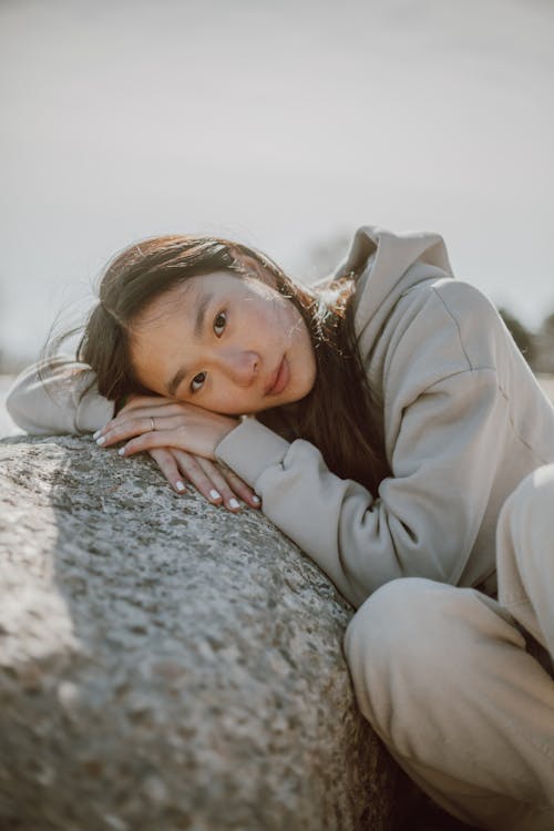Serene Asian female with long dark hair wearing casual clothes leaning on boulder and looking at camera
