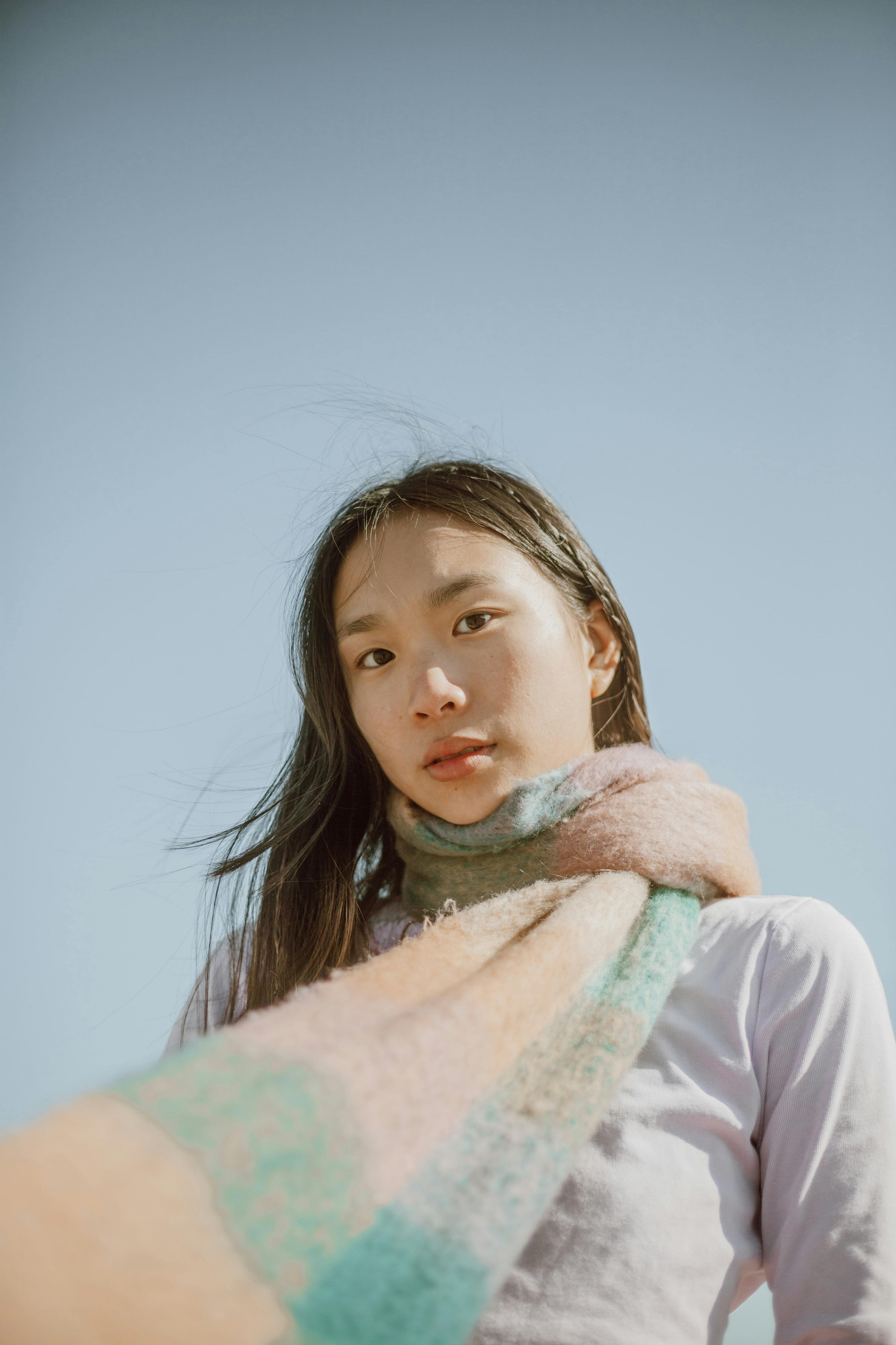 Low angle of Asian female with long dark hair wearing warm scarf standing against blue sky and looking at camera