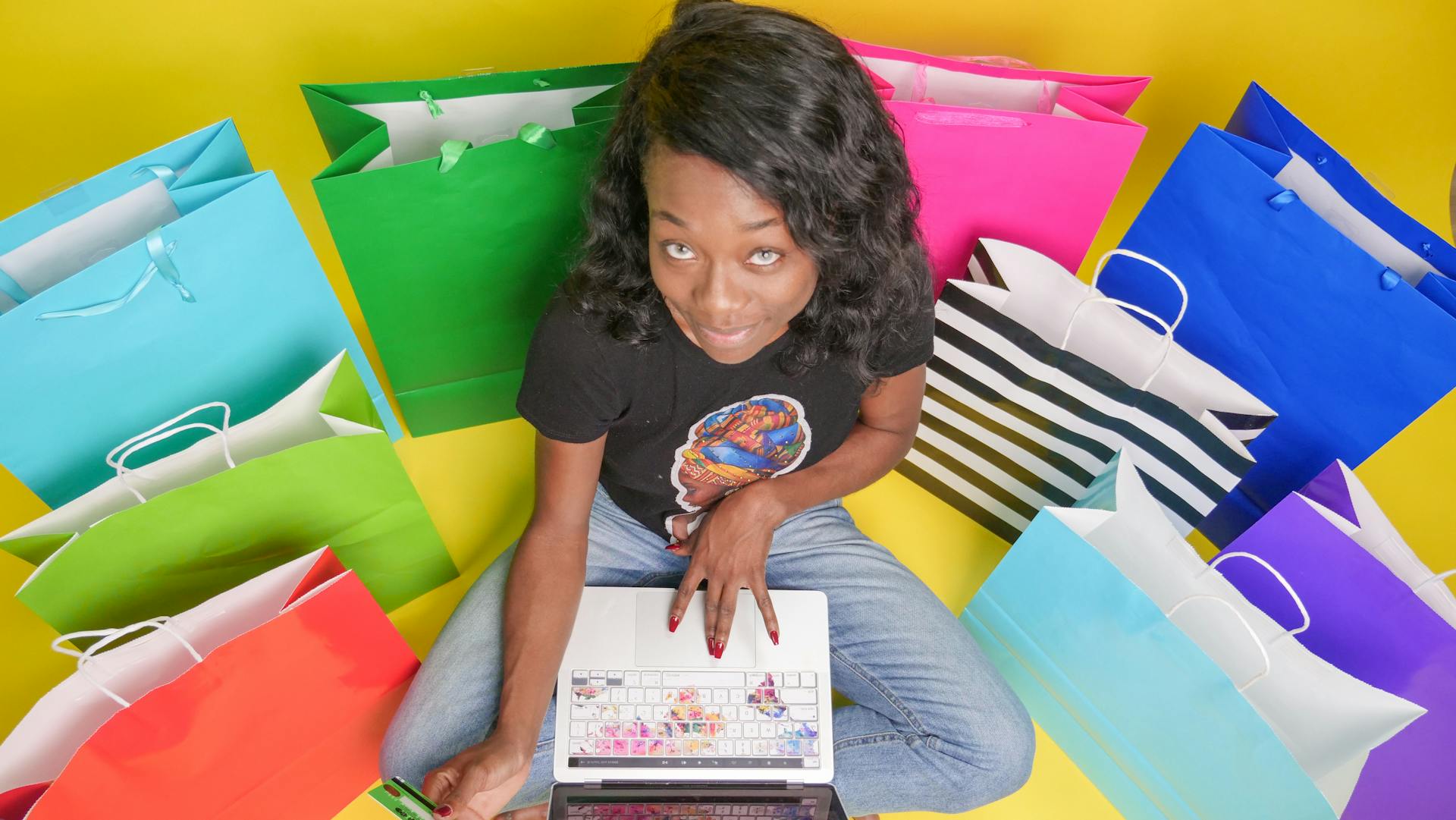 A woman browsing on her laptop surrounded by colorful shopping bags, illustrating online shopping.