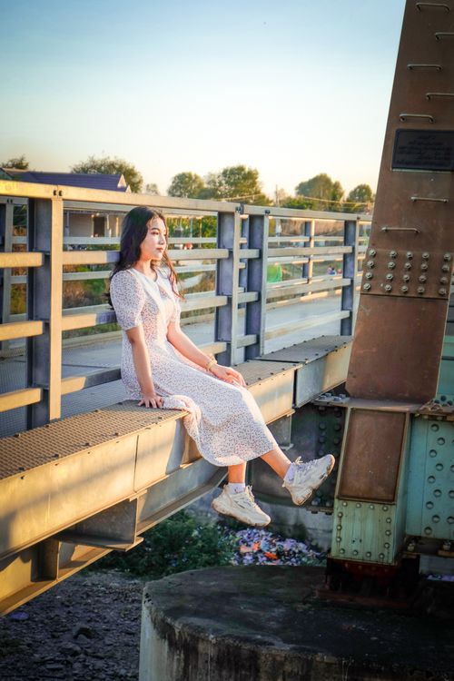 Free Young woman in summer dress sits on a bridge during a golden hour sunset. Stock Photo