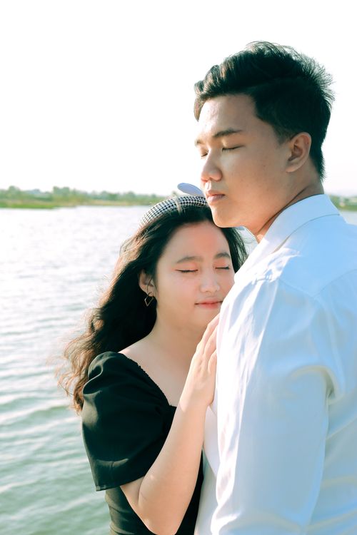 Free A couple embraces by a serene lake, both with closed eyes, enjoying a peaceful moment together. Stock Photo