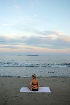 Back view of unrecognizable young female traveler with blond hair in bikini resting on sandy beach in front of wavy sea against sunset sky