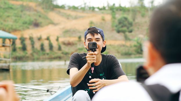 A young man smiles while holding a camera, sitting outdoors on a boat, capturing the scenic view.