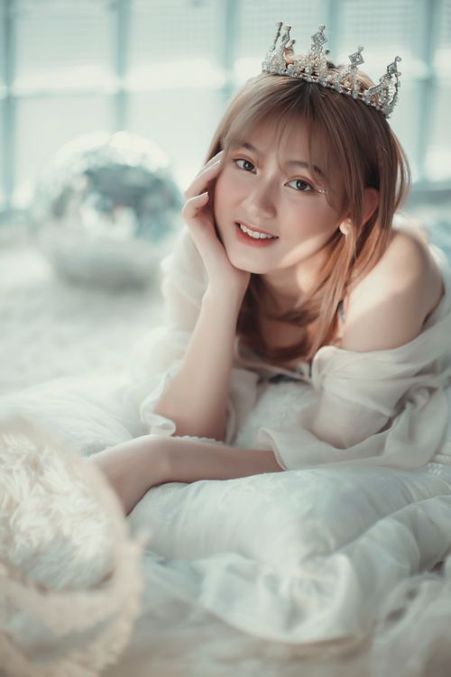 Free Beautiful Asian woman in white dress smiles while lying on a cozy bed with a sparkling tiara. Stock Photo