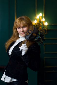 Charming woman in a black blazer holding a masquerade mask in an elegant indoor setting.
