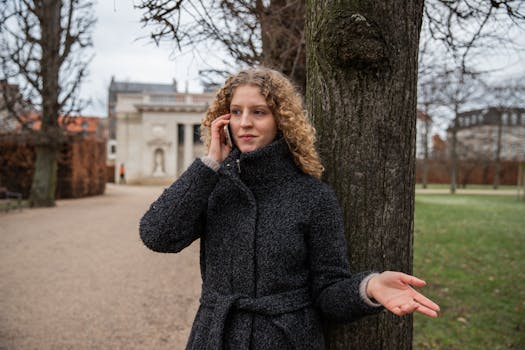 A woman in a coat talks on her phone against a tree in a Copenhagen park.