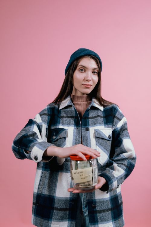 Free A woman in a plaid shirt and beanie holds a glass jar labeled savings against a pink background. Stock Photo