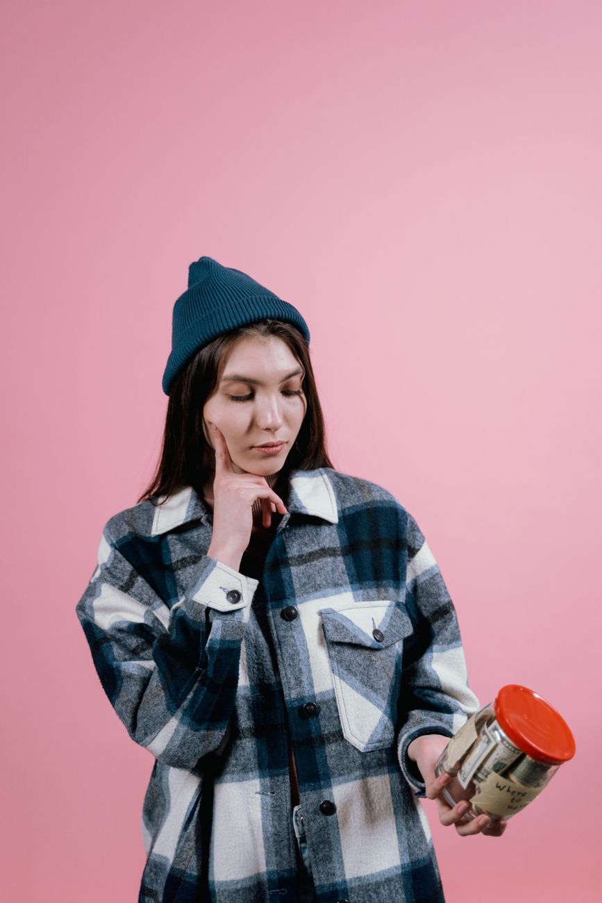 Used iPhone 15: A Smart Buy or a Gamble? 2 Thoughtful woman in beanie holding a savings jar, contemplating financial decisions, in front of a pink background.