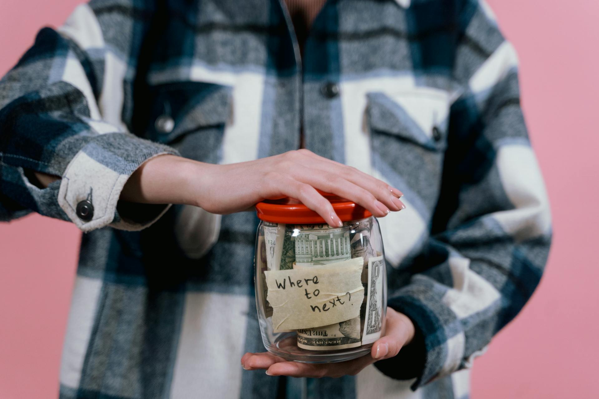 Close-up of a woman holding a savings jar labeled 'Where to next?' filled with US dollar bills.