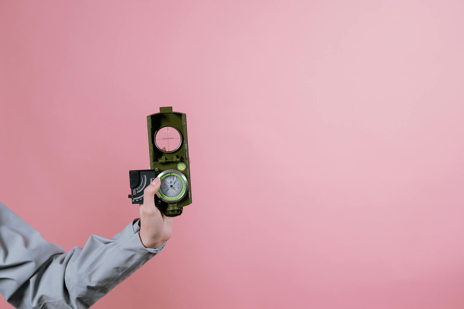 A person holds a military compass against a pink background, symbolizing navigation and direction.