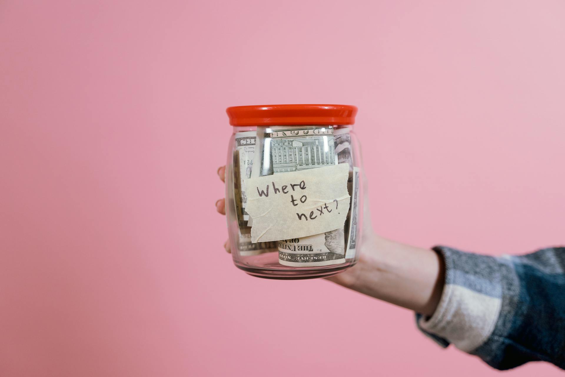 A hand holding a glass jar filled with dollar bills labeled 'Where to next?' against a pink background, symbolizing savings and future planning.