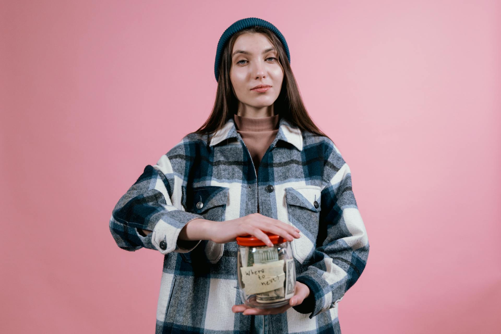 Woman wearing plaid shirt and beanie holds a money jar labeled 'where to next?' against a pink studio background.