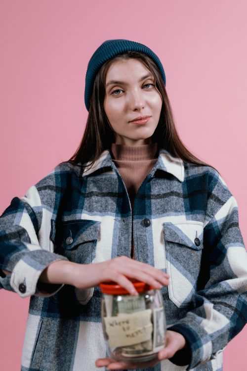 Free Portrait of a stylish woman in a beanie holding a glass jar, shot against a pink background in a studio setting. Stock Photo