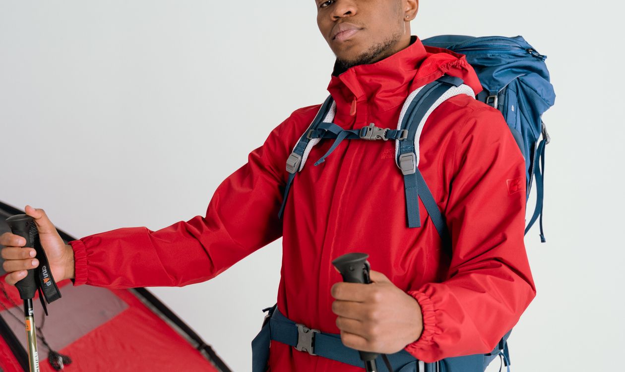 Hiker in a red windbreaker holding a pair of trekking poles at his sides, standing next to a pitched tent, illustrating correct trekking pole fit and length for hiking