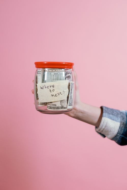 Free Close-up of a hand holding a savings jar filled with money against a pink backdrop. Stock Photo